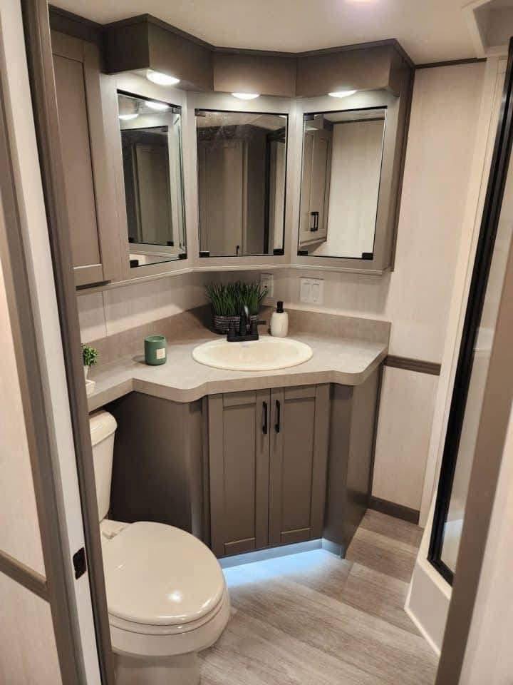 Modern bathroom with beige tones, featuring a vanity with mirrors, sink, and gray cabinets. Includes a toilet, potted plants, and soft lighting.