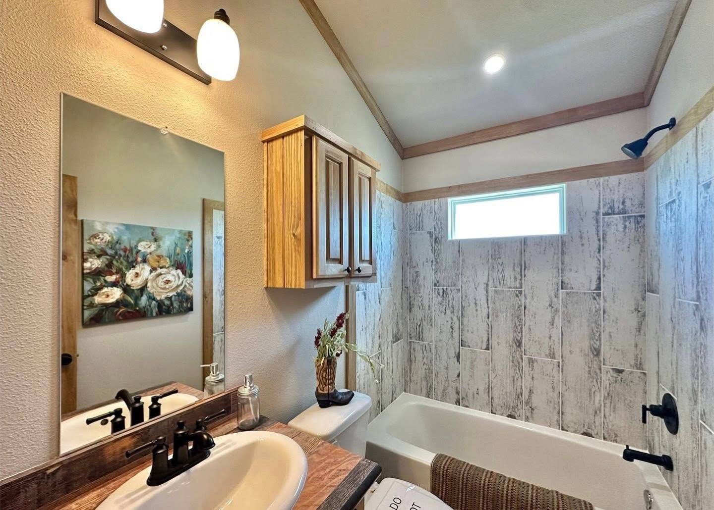 A cozy bathroom features a wooden vanity with a white sink and black fixtures, rustic wood cabinets, a floral painting, textured wall tiles, and a bathtub.