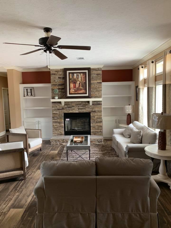 Cozy living room with beige sofas, stone fireplace, and artwork above. A ceiling fan, wooden floor, and bookshelves create a warm, inviting atmosphere.