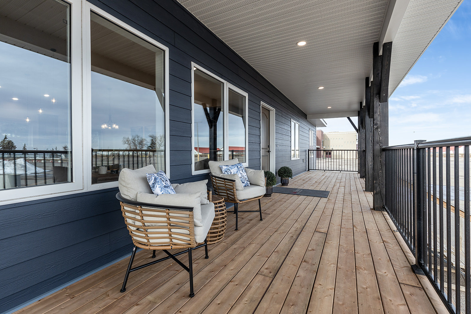 Spacious wooden deck with navy siding, featuring two wicker chairs with cushions and pillows. Overhead lighting creates a serene, inviting atmosphere.