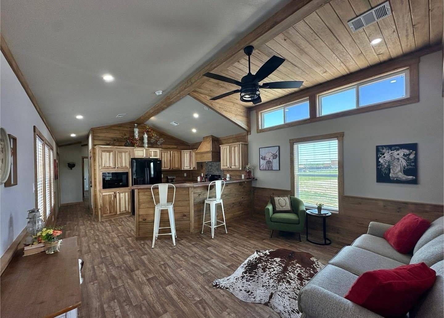 Spacious, rustic living room with wooden floors and ceiling. Cozy sofa with red cushions, animal print rug, and modern kitchen with bar stools.