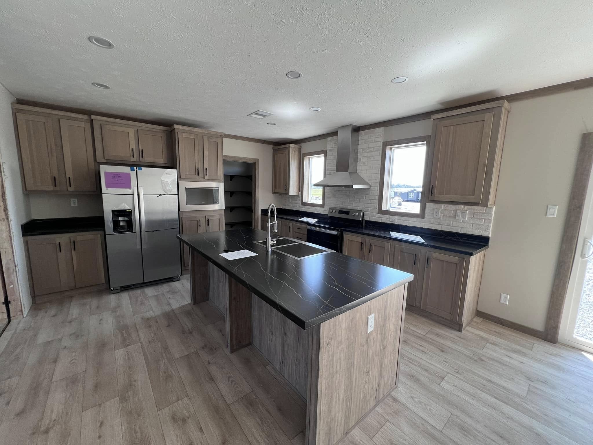 Modern kitchen with wood cabinetry and black marble countertops. Central island has a built-in sink. Stainless steel appliances; two windows provide natural light.
