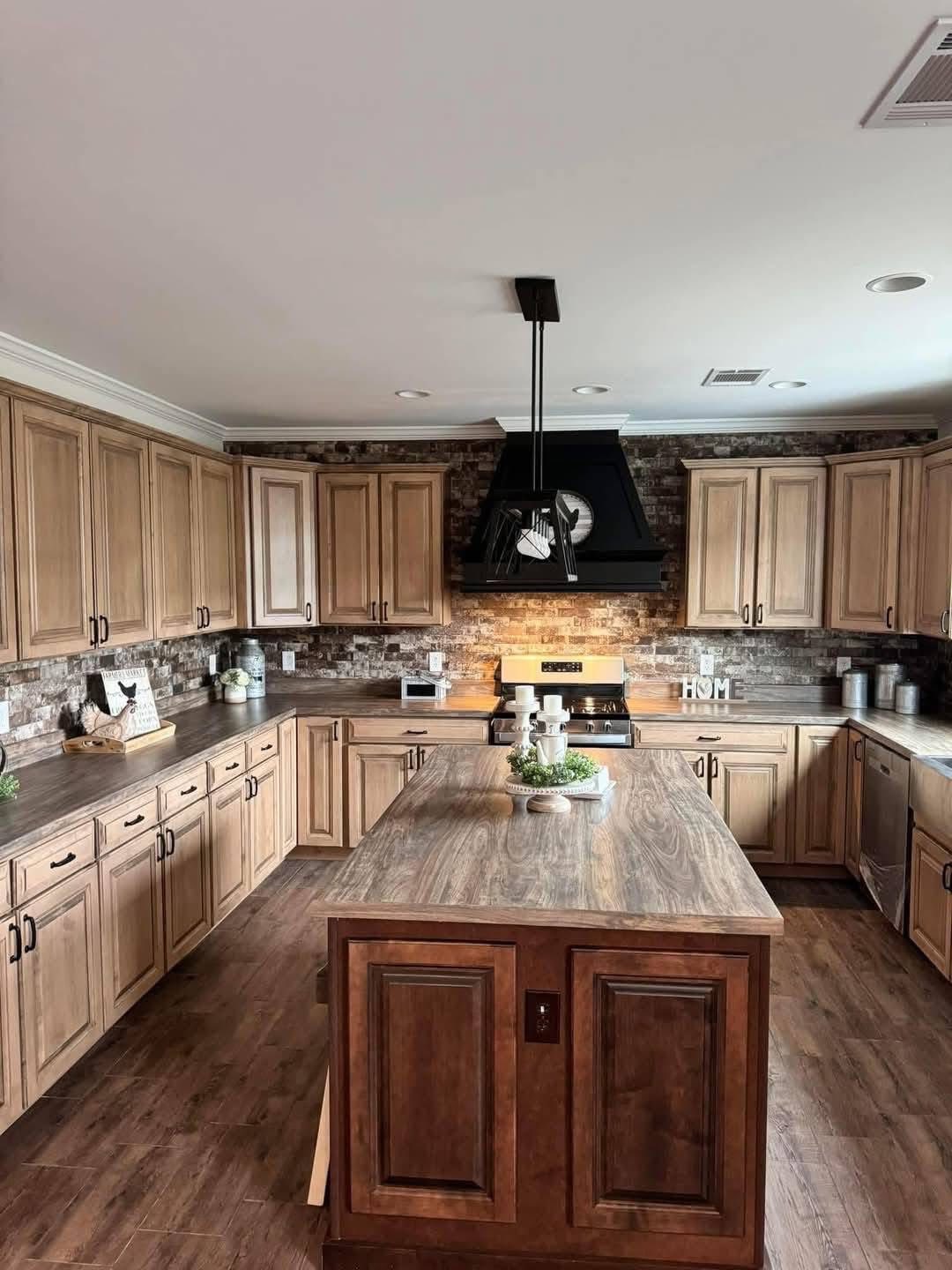 A warm, rustic kitchen with wooden cabinets and a large island. Brick backsplash, black stove hood, and ambient lighting create a cozy atmosphere.