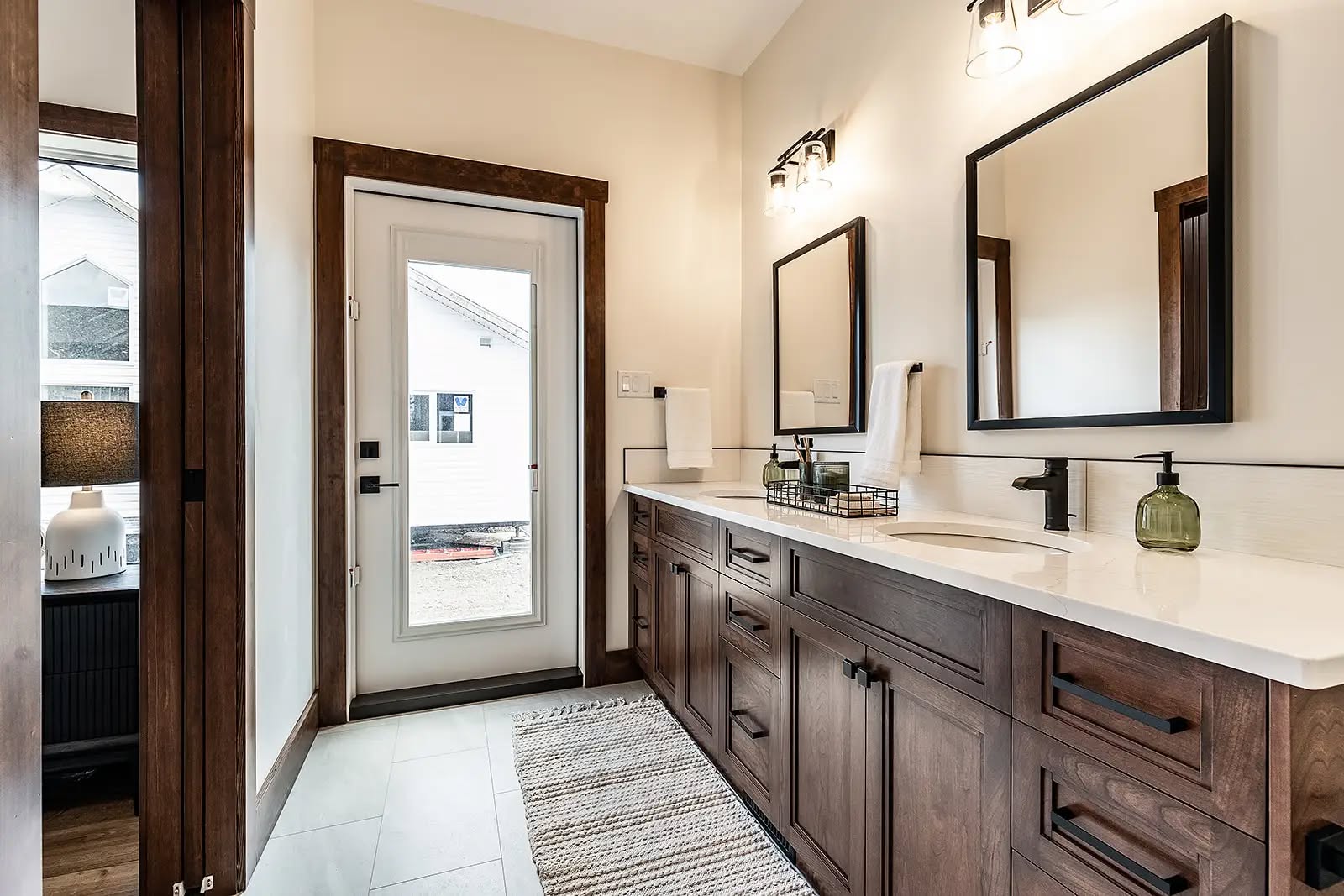 Bright bathroom with large mirror, dual sinks, and dark wood cabinets. A glass door leads outside. Neutral tones create a modern, cozy feel.