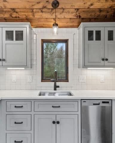 Gray kitchen with shaker cabinets, a farmhouse sink, and a stainless steel dishwasher. A window offers a view of trees, under a wooden ceiling with a pendant light.