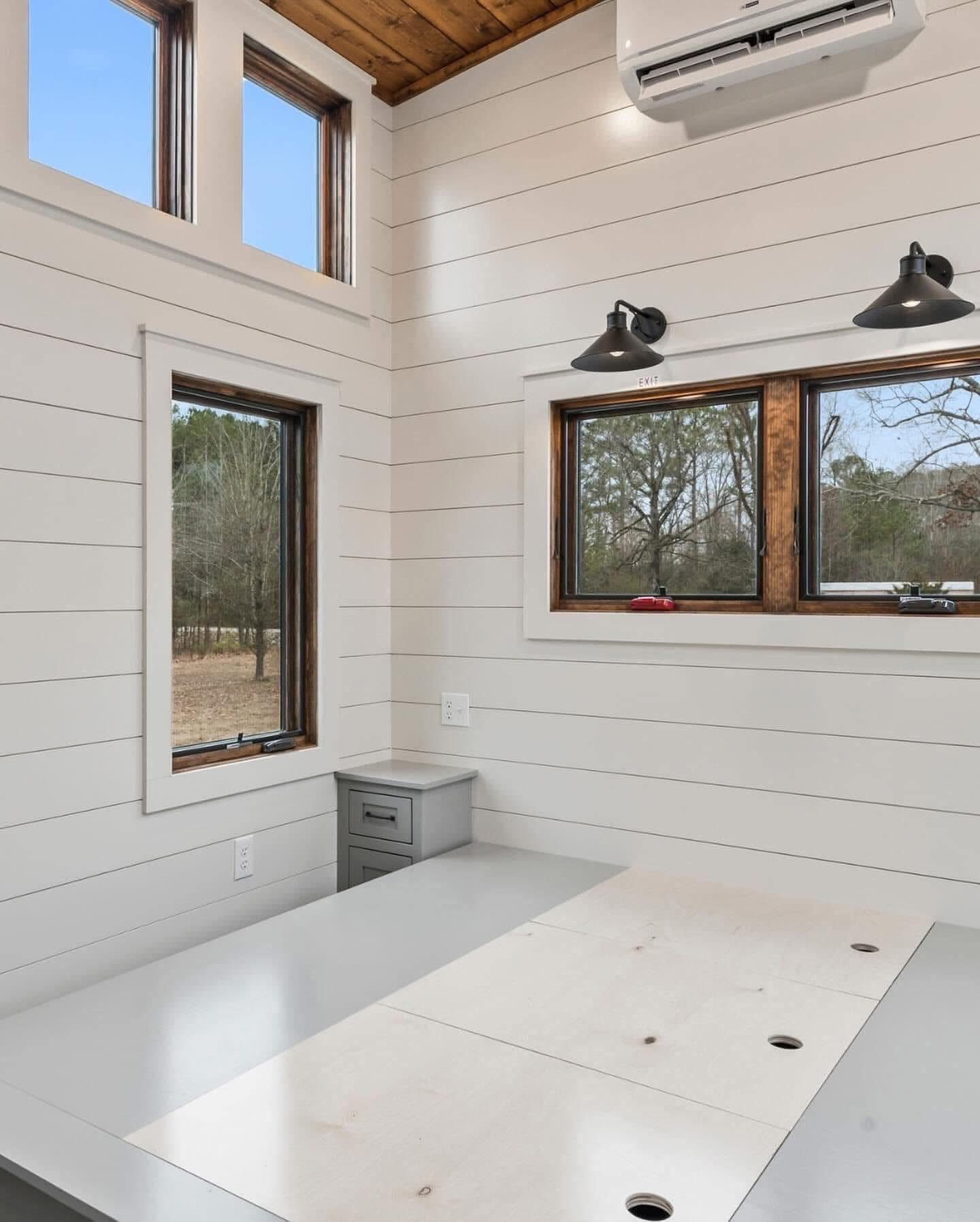 Minimalist room with white shiplap walls and large windows, featuring a light gray desk and small gray drawers. Warm lighting creates a cozy atmosphere.