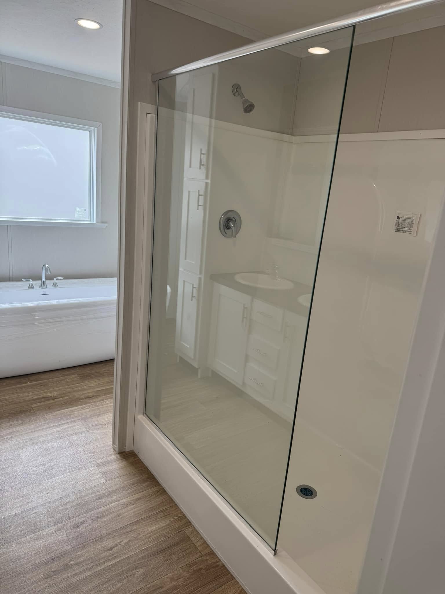 Modern bathroom with a clear glass shower door, white fixtures, and wood-style flooring. A large bathtub is by a window, creating a tranquil atmosphere.