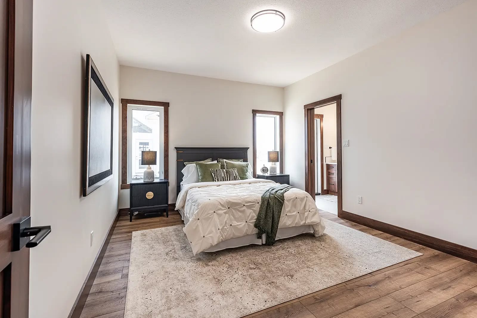A cozy bedroom features a double bed with white bedding and green pillows. Two narrow windows flank the bed, and nightstands with lamps add warmth. A soft rug lies on the wooden floor, enhancing the inviting ambiance.