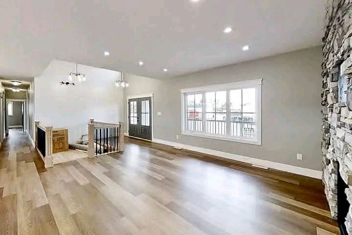 Spacious living room with wooden floors, gray walls, and a large stone fireplace. Bright natural light from a big window, modern pendant lights.
