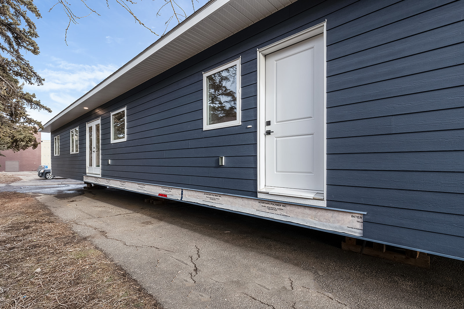 A blue manufactured home on a trailer with a white door and windows sits on pavement. Trees and a small building are visible in the background under a clear sky.
