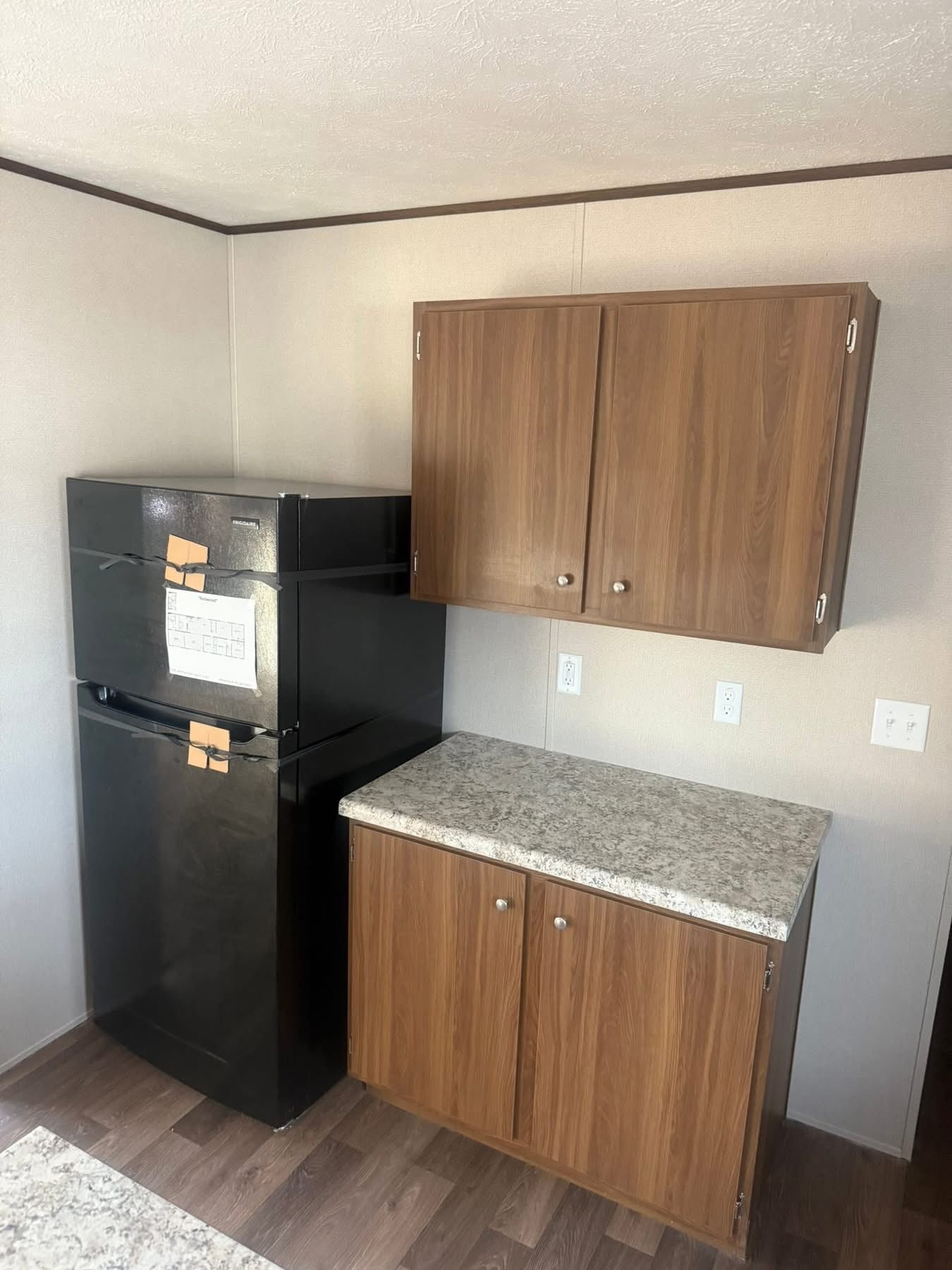 A compact kitchen corner with a black fridge, two brown cabinets, and a speckled countertop. The floor is wood, imparting a clean, minimal look.