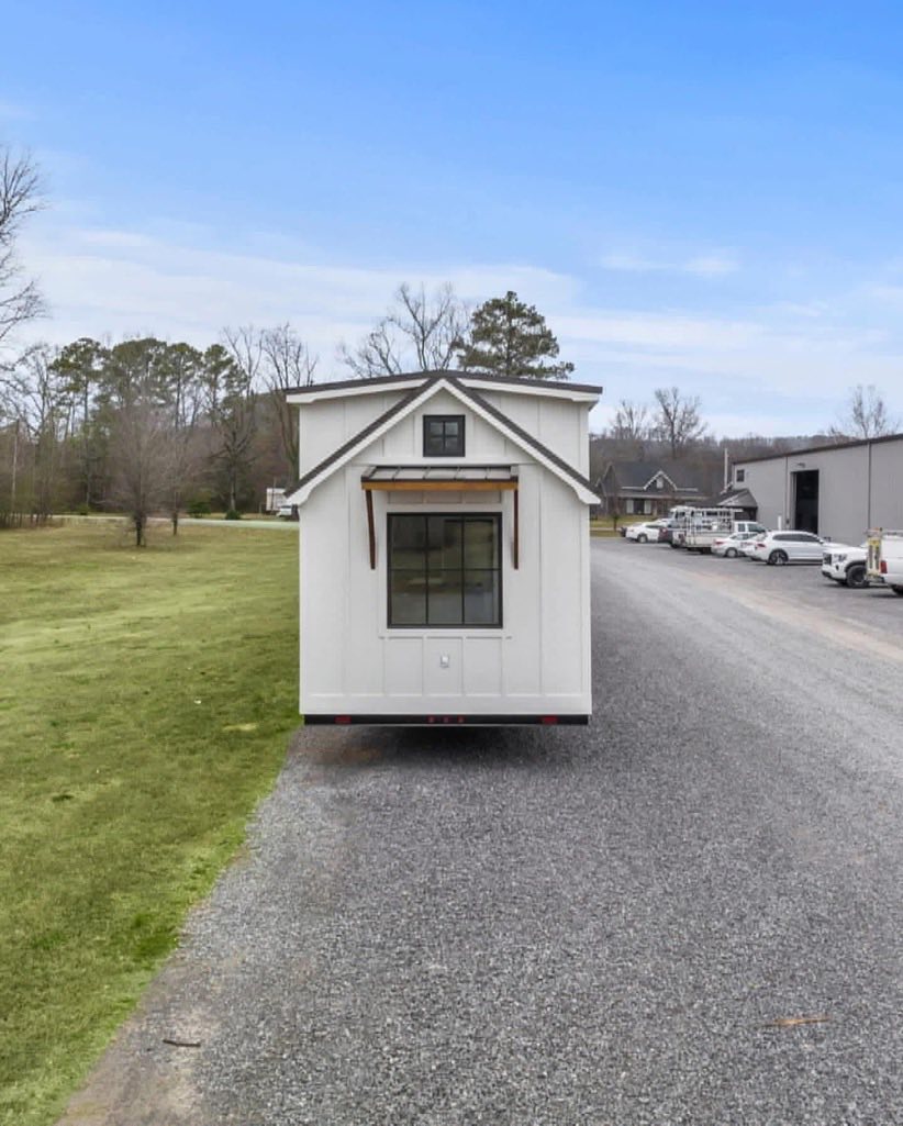 Tiny white house on wheels parked on a gravel road, surrounded by green grass and bare trees under a clear blue sky, conveying simplicity and mobility.