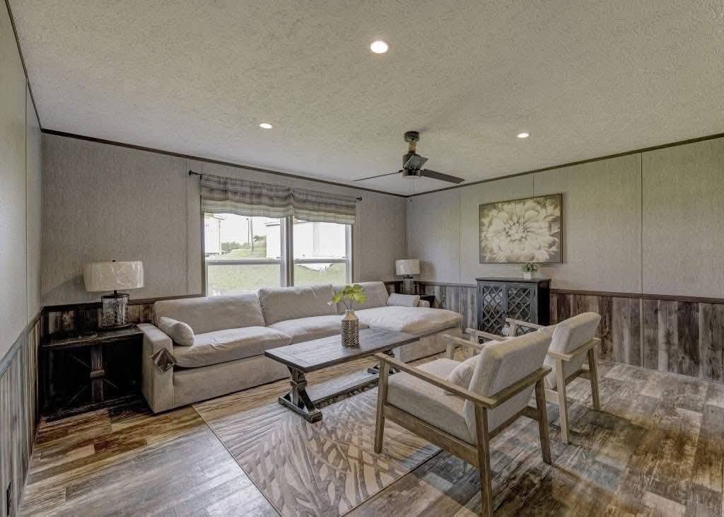 Cozy living room with gray sofas, wooden coffee table, patterned rug, and two armchairs. Large floral artwork and windows with natural light enhance the space.