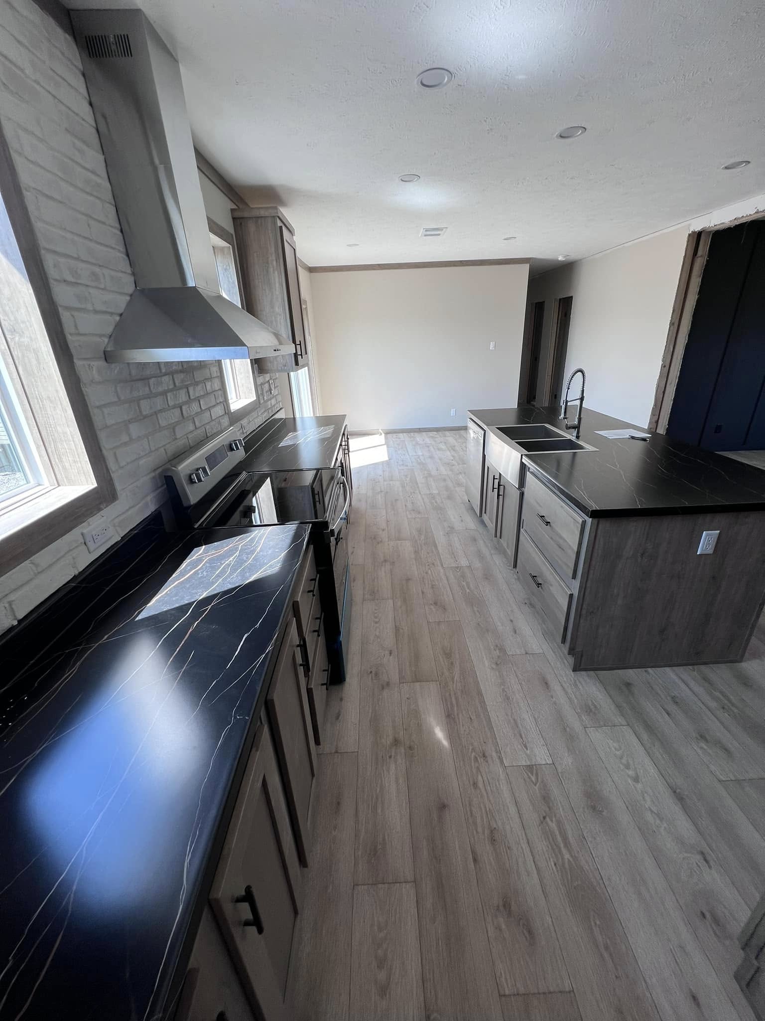 Modern kitchen with sleek black marble countertops and wooden cabinets. Includes a stainless steel stove, vent hood, and a central island sink on light wood flooring. Bright natural light from windows.
