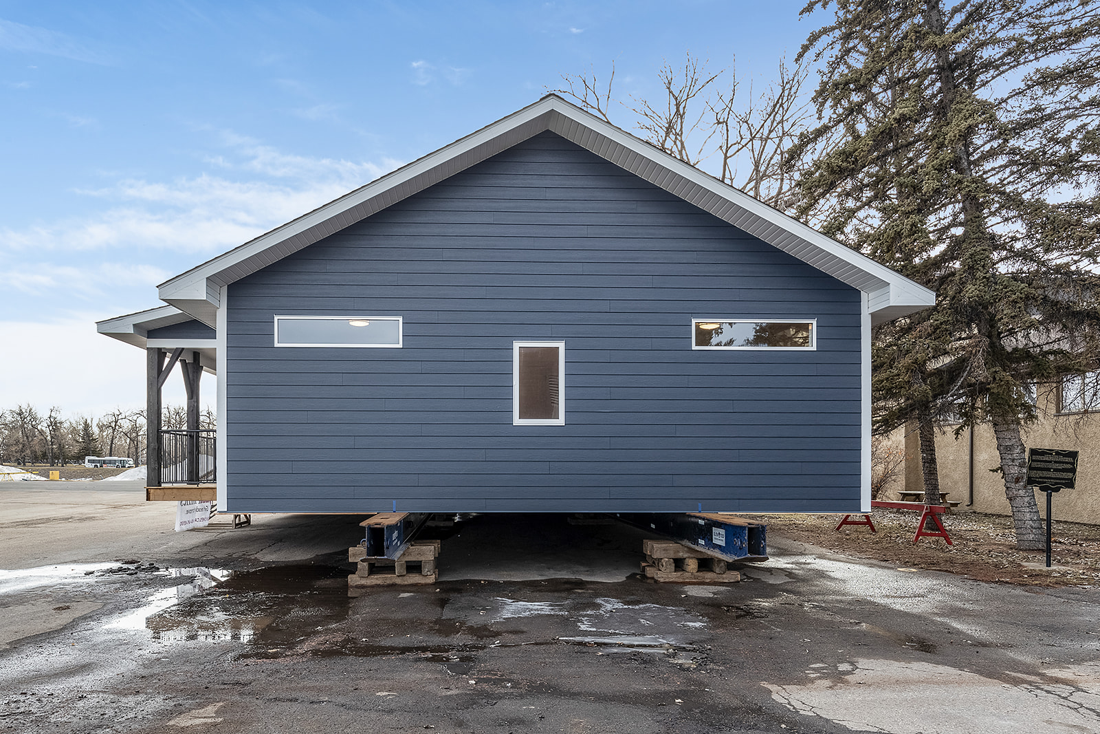 Front view of a blue modular home on pallets, placed on an asphalt surface. The sky is partly cloudy, and bare trees stand in the background.