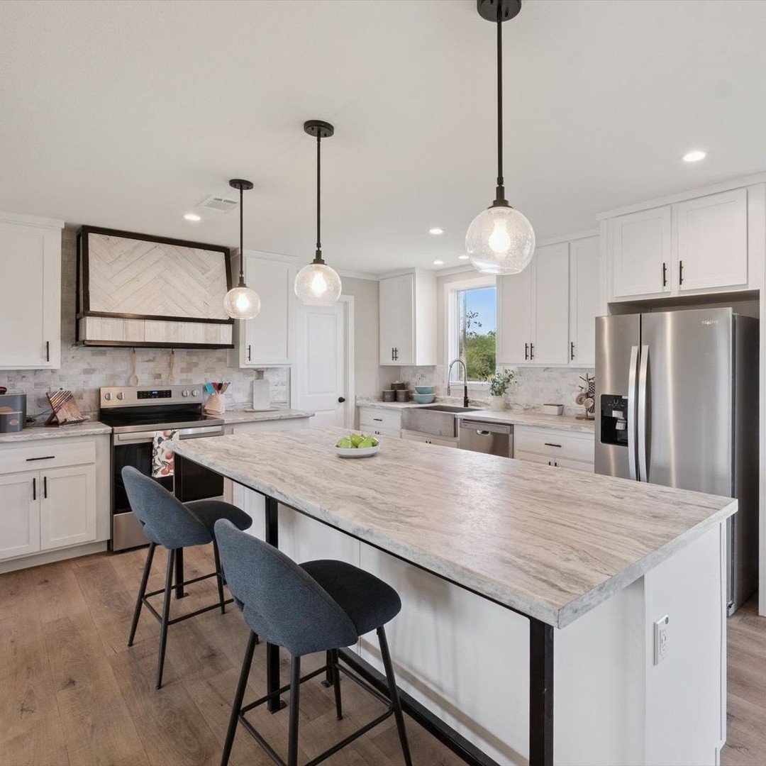 Modern kitchen with white cabinets, a marble island, and three pendant lights. Stainless appliances and two blue chairs create a sleek, inviting space.