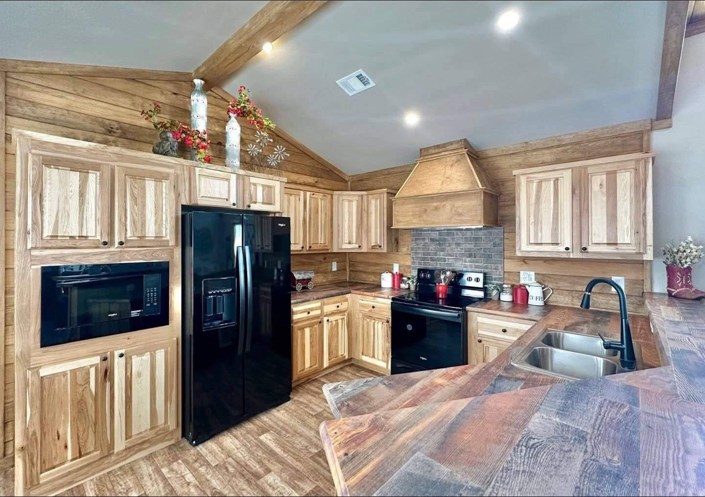 Rustic kitchen with wooden cabinets, black appliances, and a modern sink. Decor includes red accents and plants, creating a cozy, warm atmosphere.