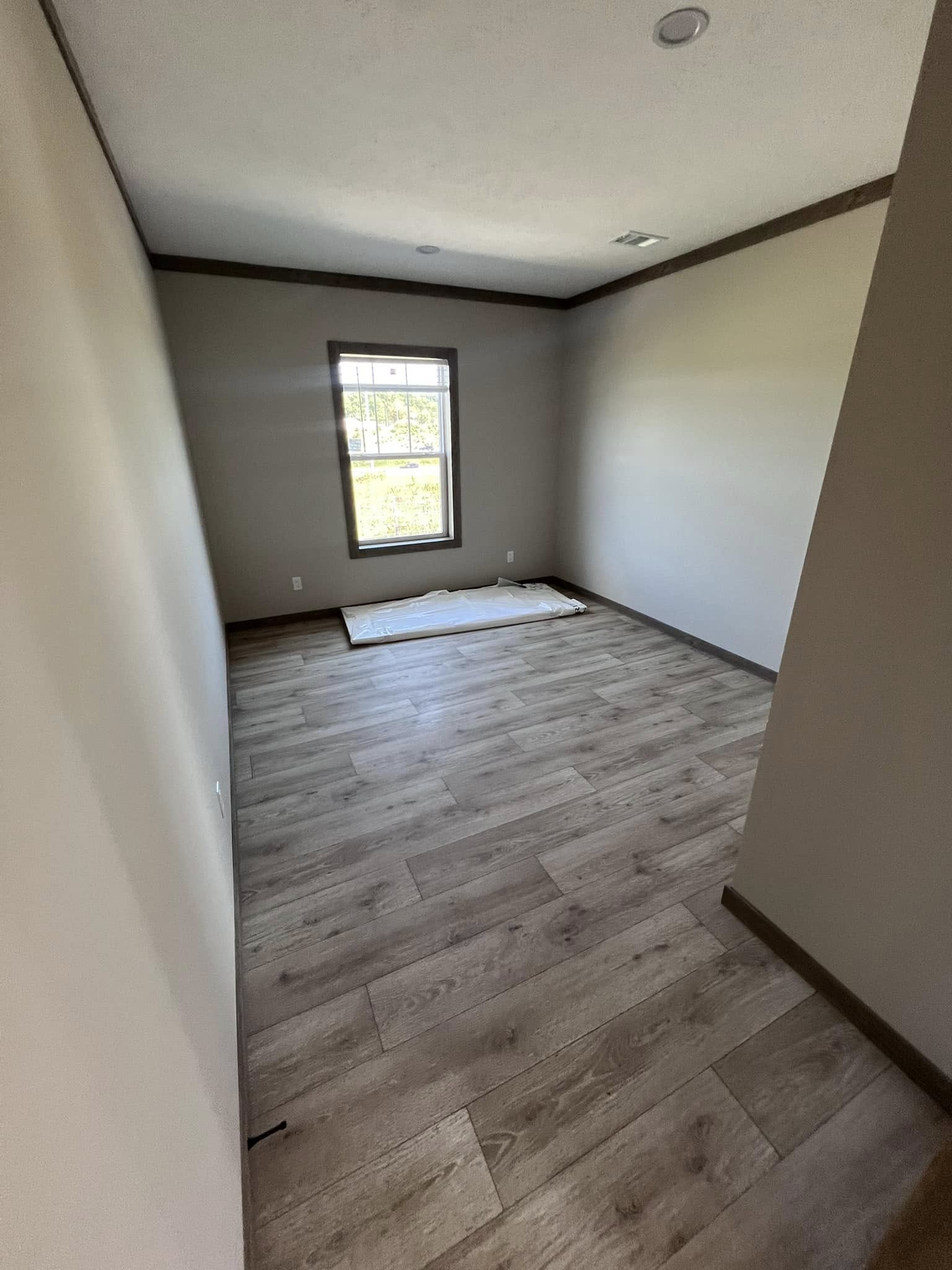 A narrow, empty room with light wood flooring and beige walls. A window with natural light and a view of greenery brings warmth to the otherwise bare space.