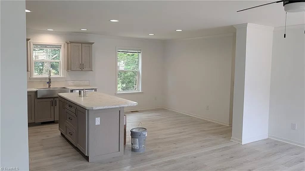 A modern kitchen with light wood floors, beige cabinets, and a marble-topped island. Natural light enters through windows, creating a fresh, clean look.