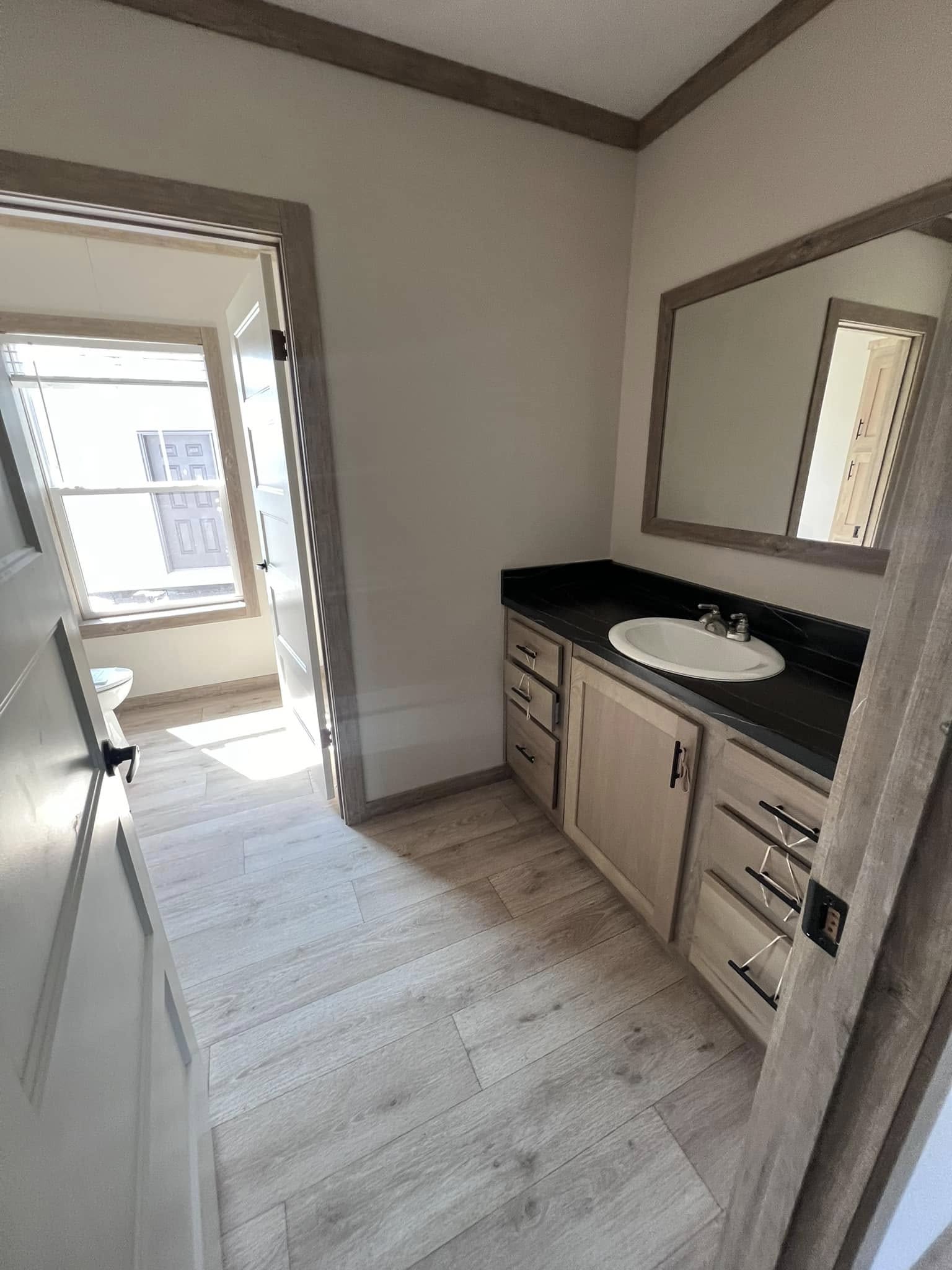 A small bathroom with light wood flooring and cabinetry. A white sink with a black countertop is below a framed mirror. Sunlight streams through a window.