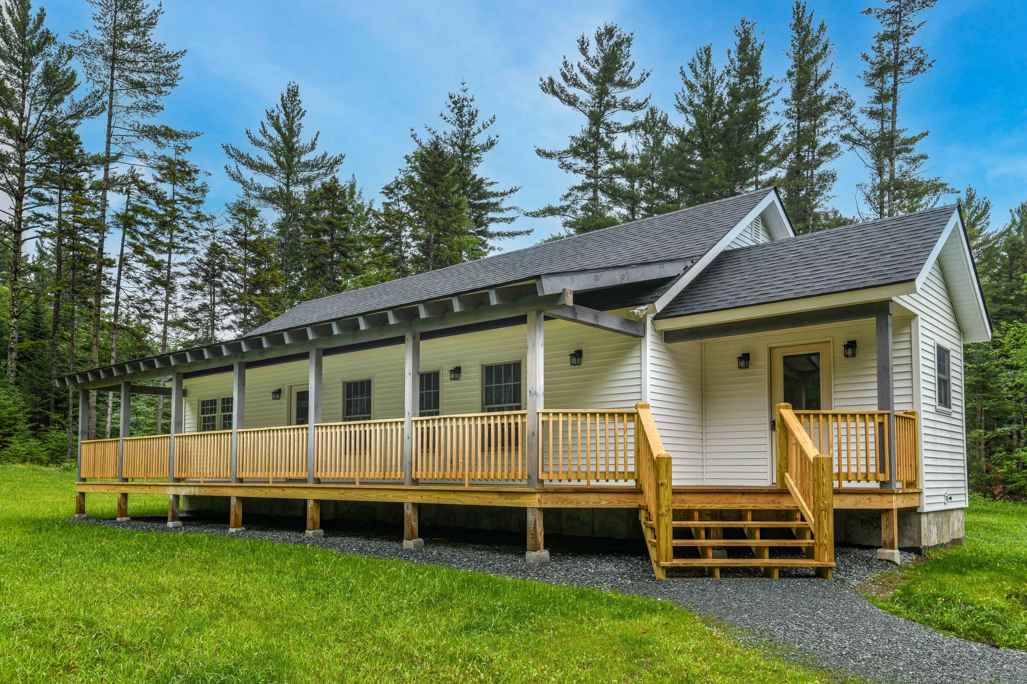 A cozy white cabin with a wooden porch set against a backdrop of tall pine trees. The structure is surrounded by lush green grass, evoking a serene, rustic charm.