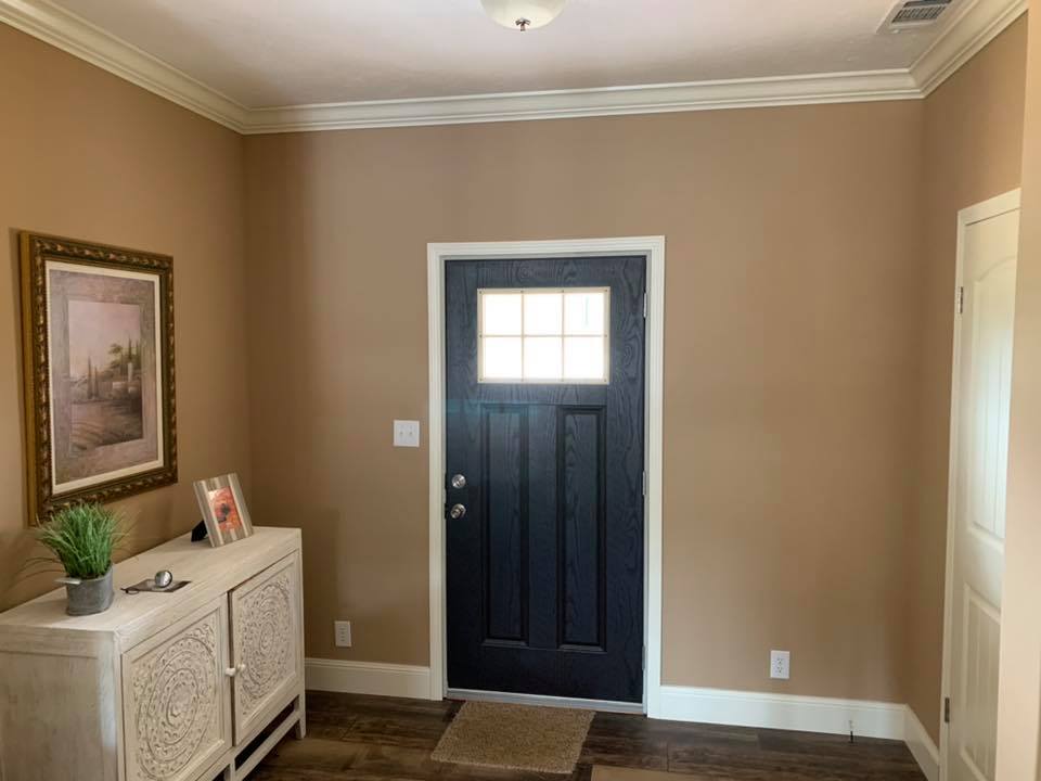 Entryway with a black door featuring a grid window. Tan walls, white trim, a decorative cabinet with a plant, and landscape painting create a warm, inviting atmosphere.