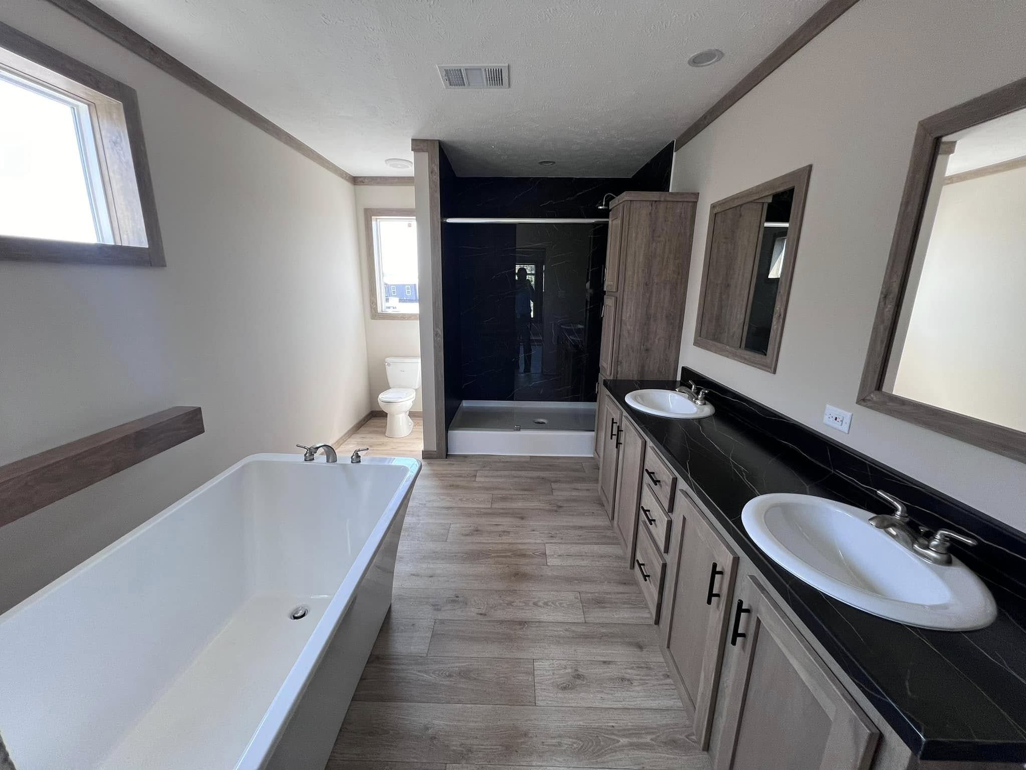 Spacious modern bathroom with a bathtub on the left, wood flooring, black-accented double sinks on the right, and a shower next to a window-lit toilet.