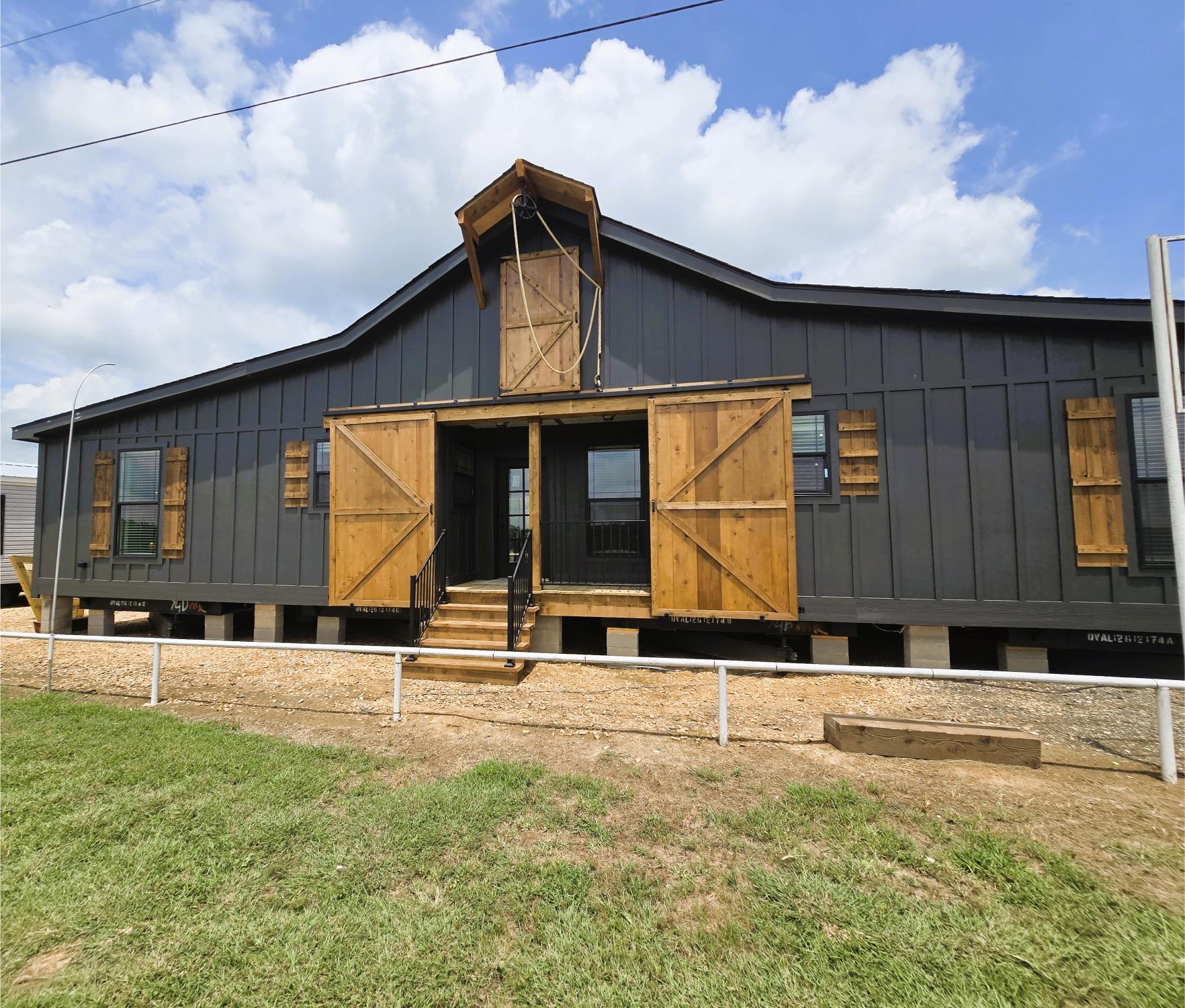 A black barn-style building with large wooden shutters and doors, elevated on stilts. Bright daytime sky with scattered clouds, conveying a rustic feel.