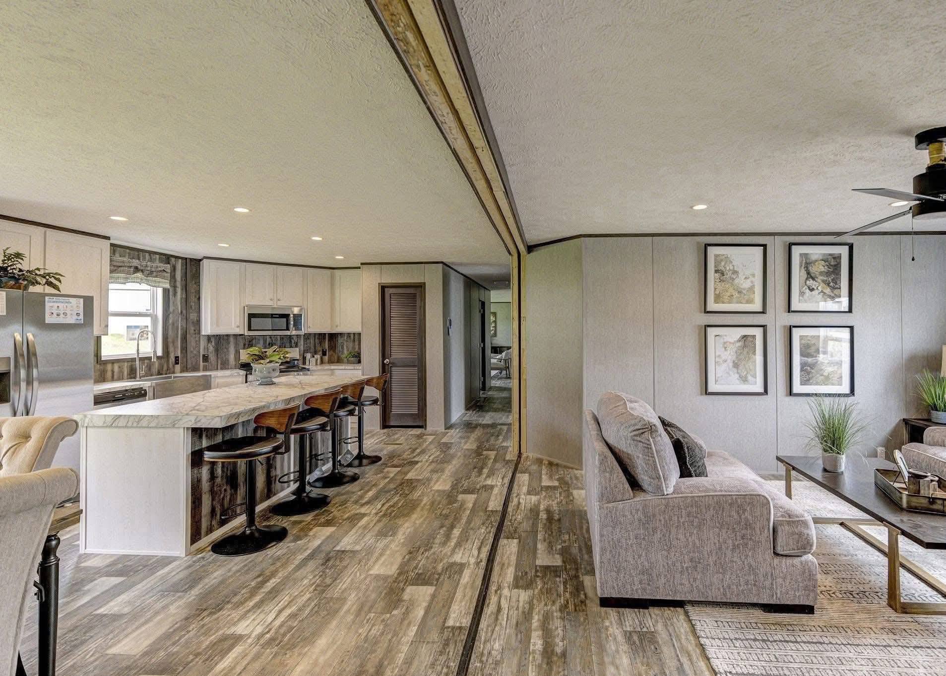 Spacious open-plan kitchen and living room with light wood floors. The kitchen has a marble island with stools, and the living area has a sofa and framed artwork.