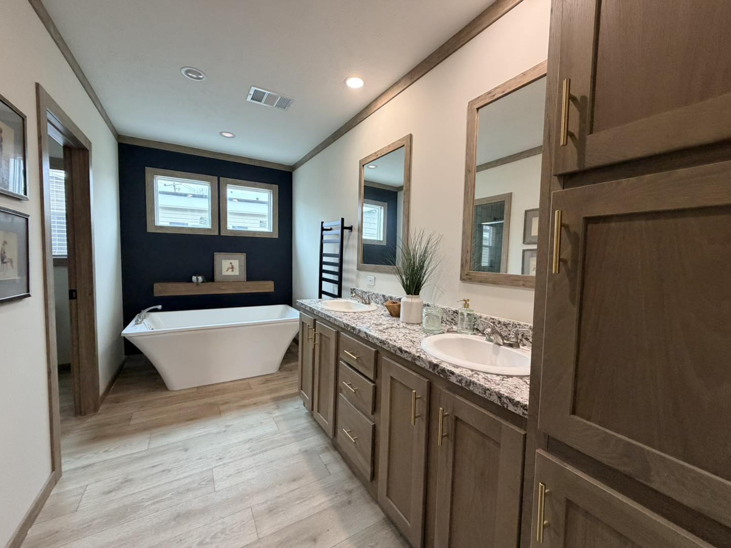 Modern bathroom with wooden cabinets, dual sinks, and a granite countertop. A standalone bathtub sits against a dark accent wall with framed windows. Warm, inviting feel.