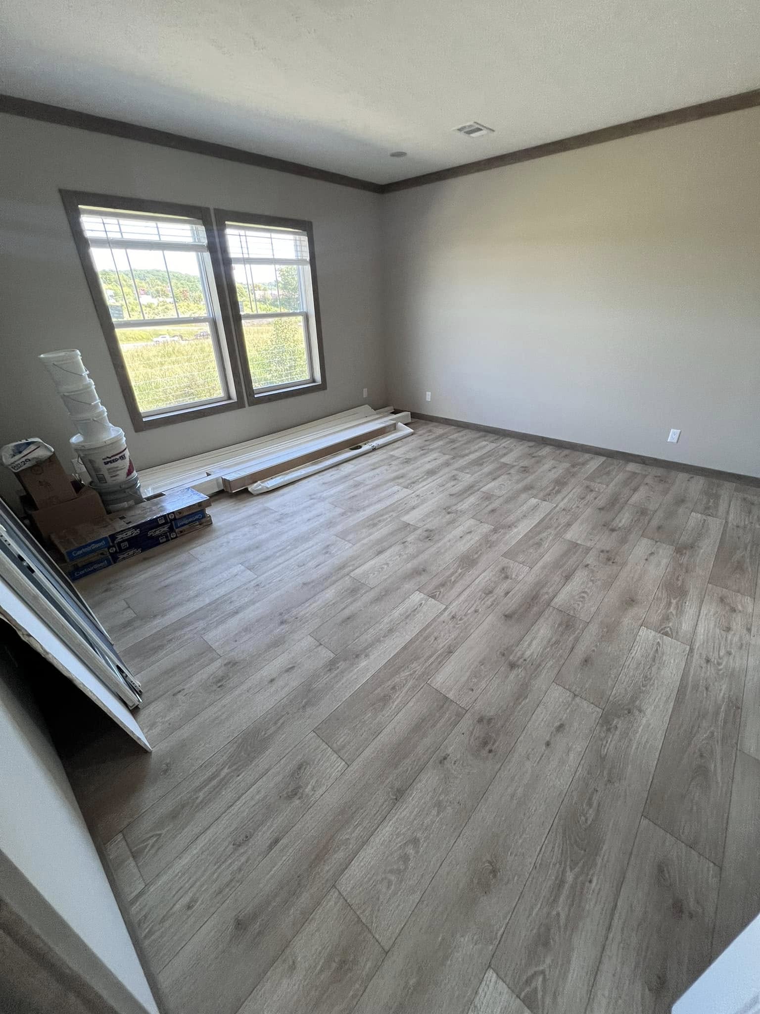 A spacious, empty room with light wood flooring and soft beige walls. Large windows on the left let in natural light. Flooring materials are stacked near the window.