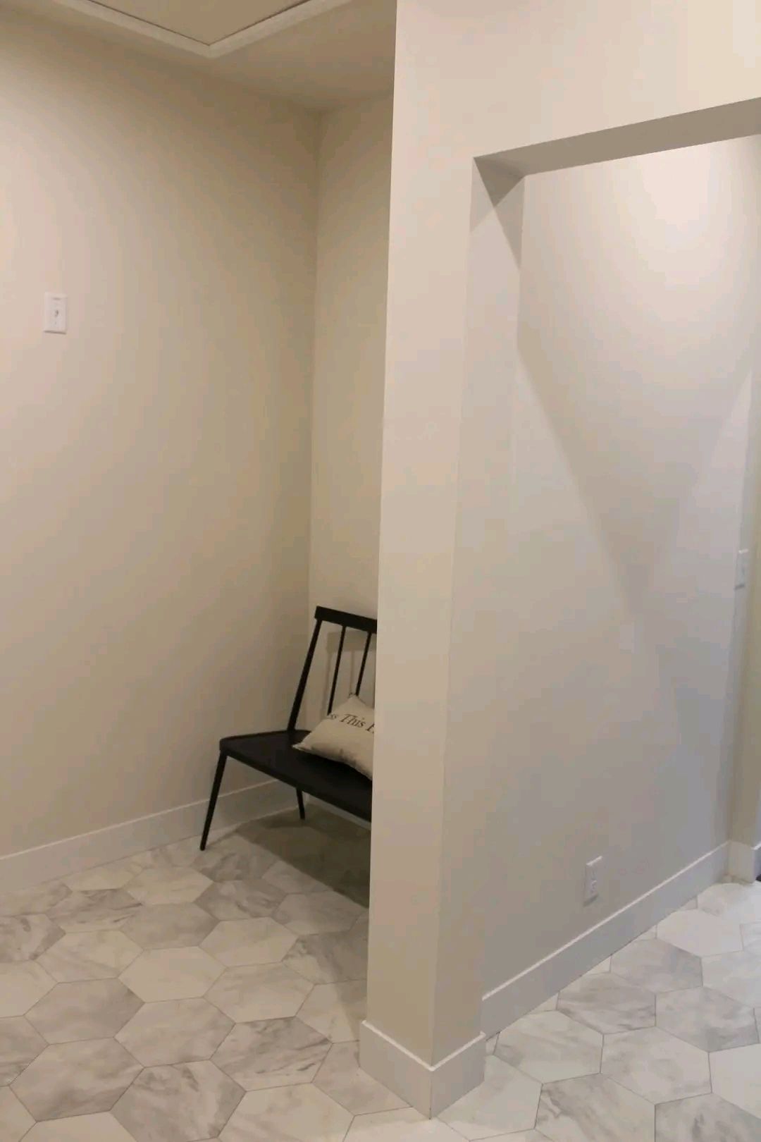 Minimalist hallway with hexagonal marble floor tiles, a black metal bench, and a cushion. The neutral walls and simple decor create a calm atmosphere.