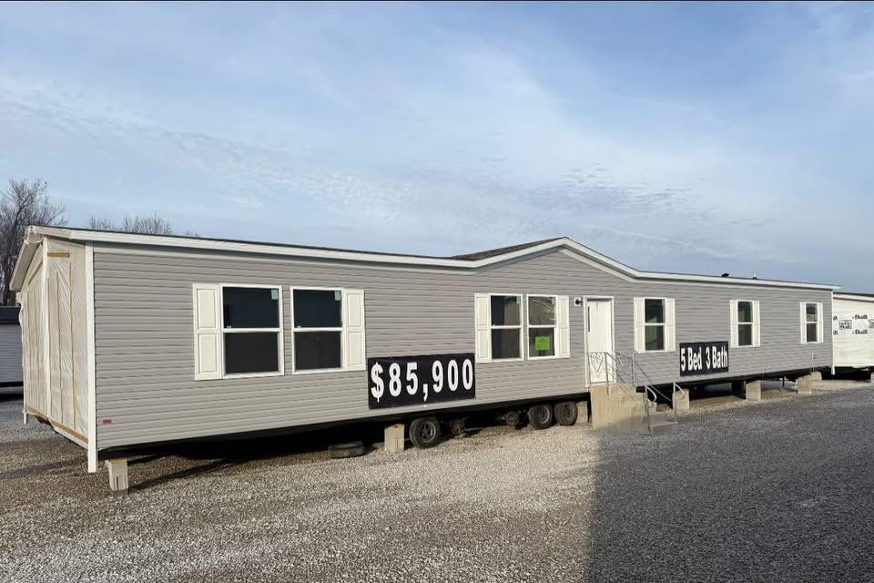 Gray mobile home with a pitched roof stands on gravel. Large signs display "$85,900" and "5 bed 3 bath" under a partly cloudy sky.