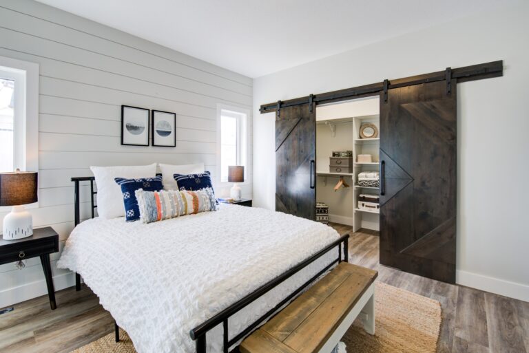Cozy bedroom with a white bed, colorful pillows, and dark wood barn doors leading to a closet. Warm lighting and shiplap walls create a rustic feel.