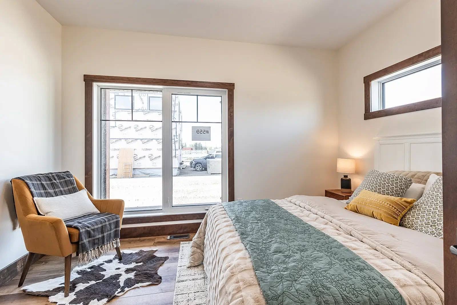 Bright bedroom with a large window, featuring a cozy bed with green quilt and golden pillows. A mustard armchair with a plaid blanket sits nearby.