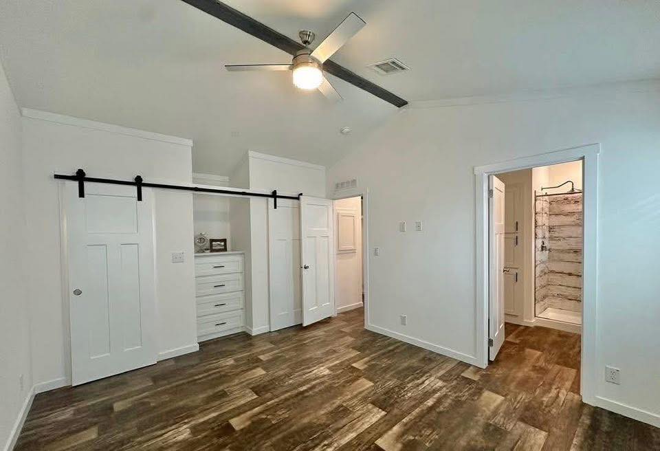 Spacious room with wooden flooring, white walls, and ceiling fan. Features sliding barn doors, built-in drawers, and an open doorway to a bathroom. Cozy and modern.