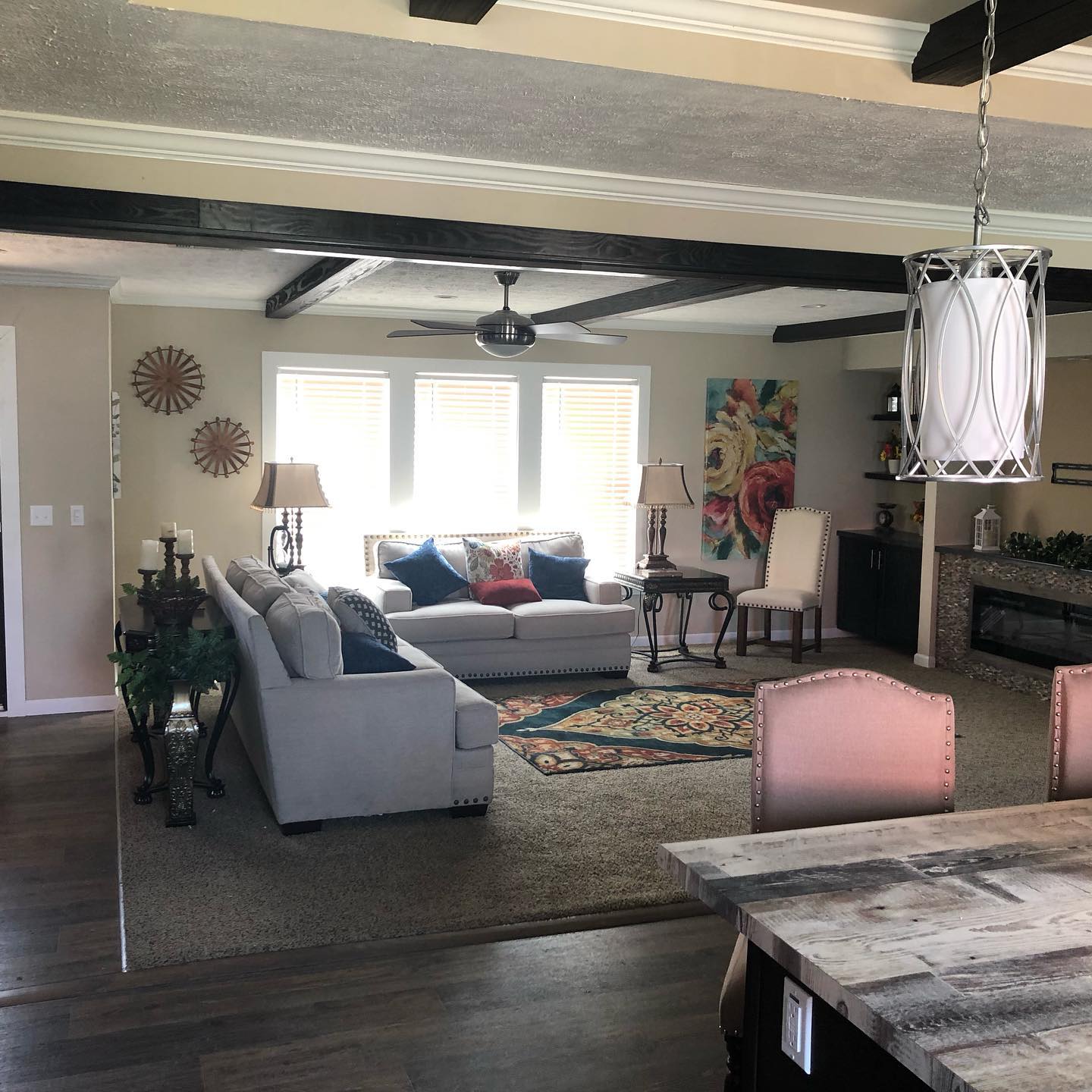 Spacious living room with modern decor, featuring a gray sofa, colorful cushions, vibrant wall art, a ceiling fan, and a rustic wooden table. Bright and inviting.
