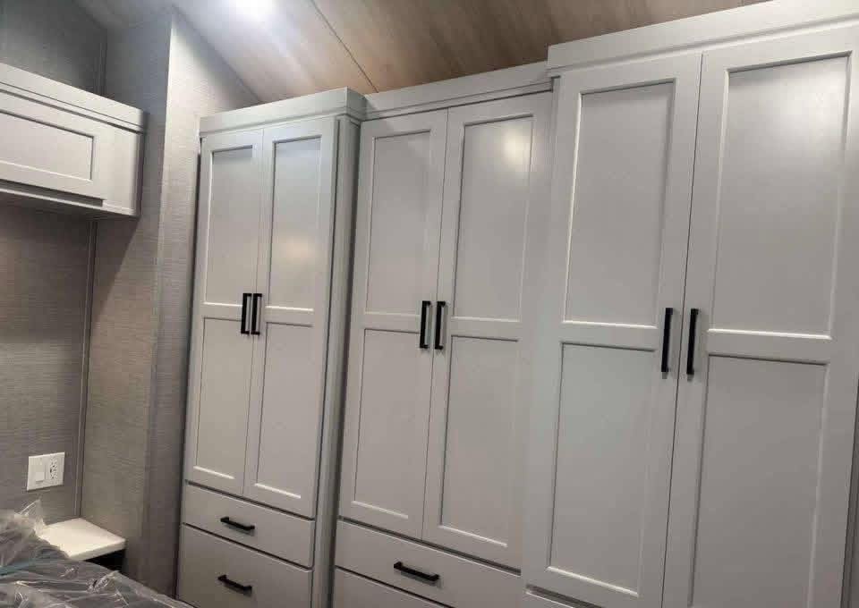 White wardrobe cabinets with black handles and clean lines, set against a light gray wall. A small countertop and electrical outlet are visible on the left.