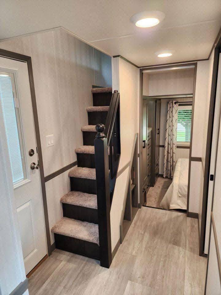 Carpeted stairs with a dark railing lead up from a small, modern hallway with light wood flooring. A cozy bedroom with a window is partially visible.