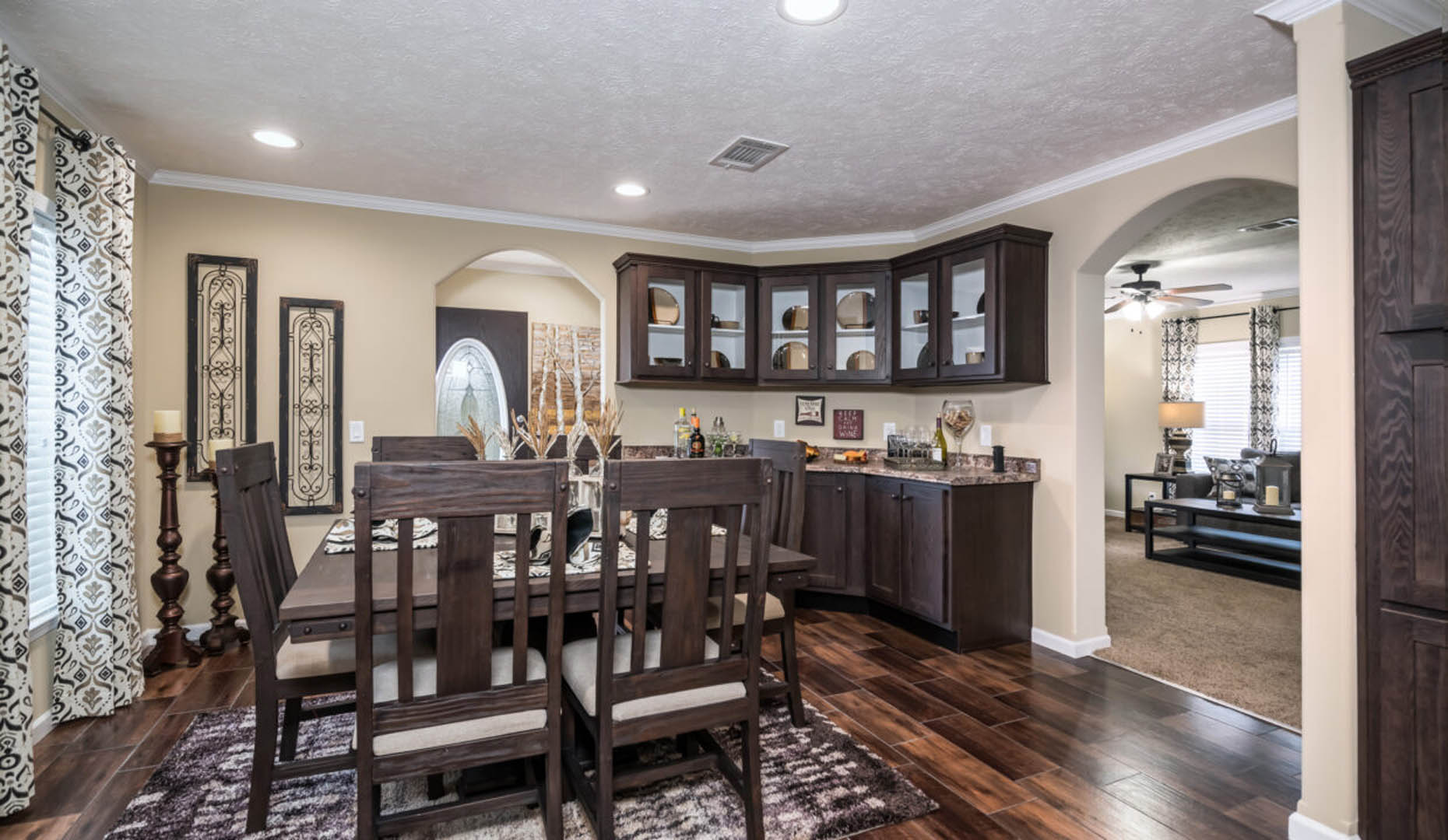 Dining room with dark wooden furniture, including a table and chairs on a patterned rug. Cabinets hold decor. Arched doorway leads to a well-lit living room.
