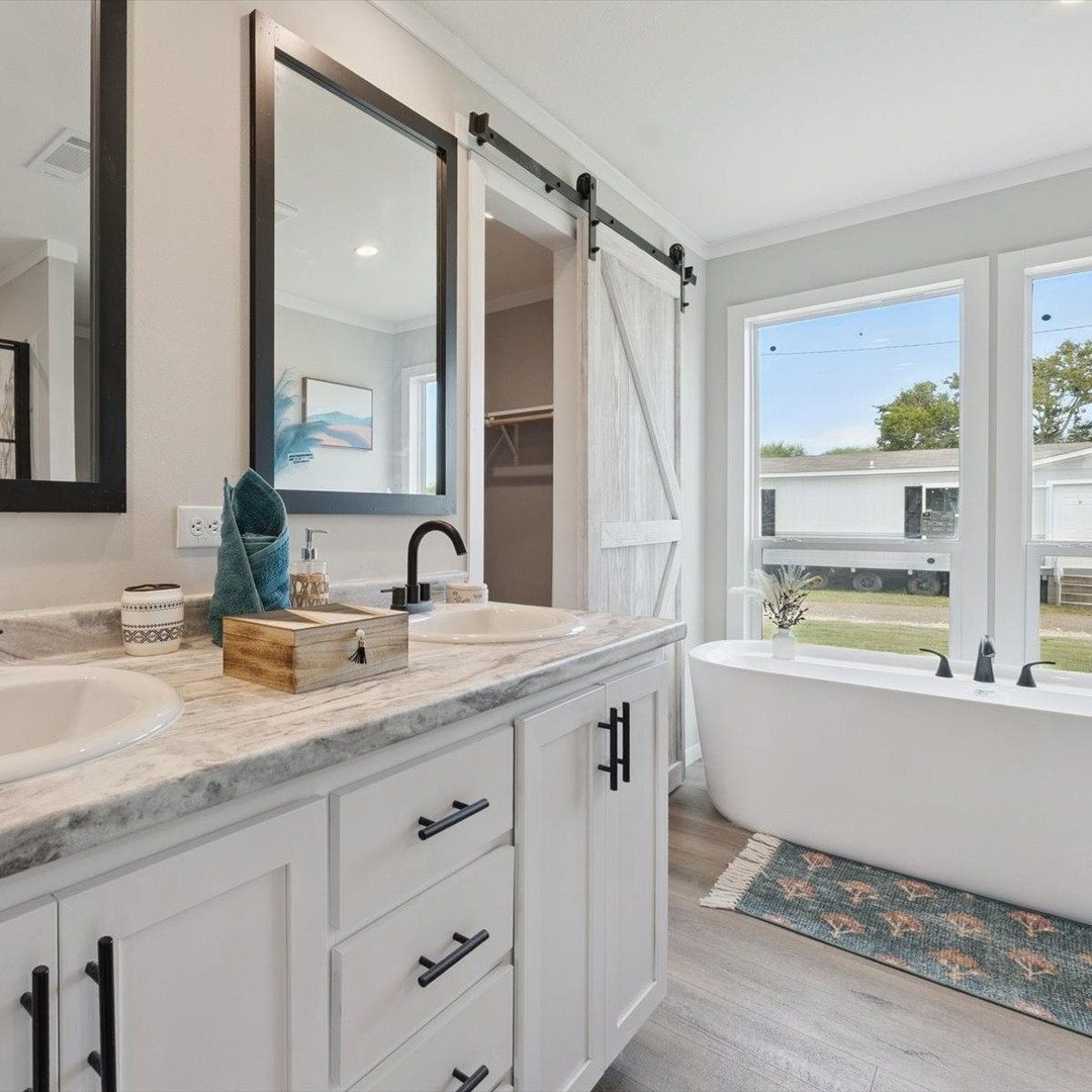 Bright bathroom with dual sinks, marble countertop, and black accents. A freestanding tub sits by large windows, next to a rustic sliding barn door.