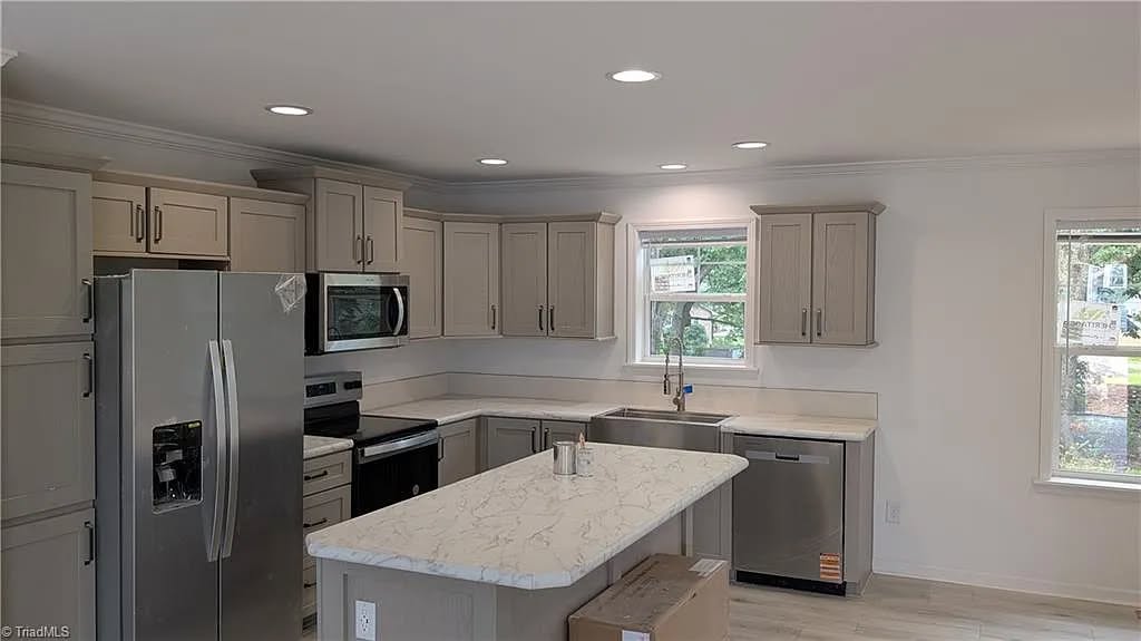 Modern kitchen with beige cabinets, stainless steel appliances, and a marble island. Bright lighting from overhead fixtures and two windows creates an open, inviting atmosphere.