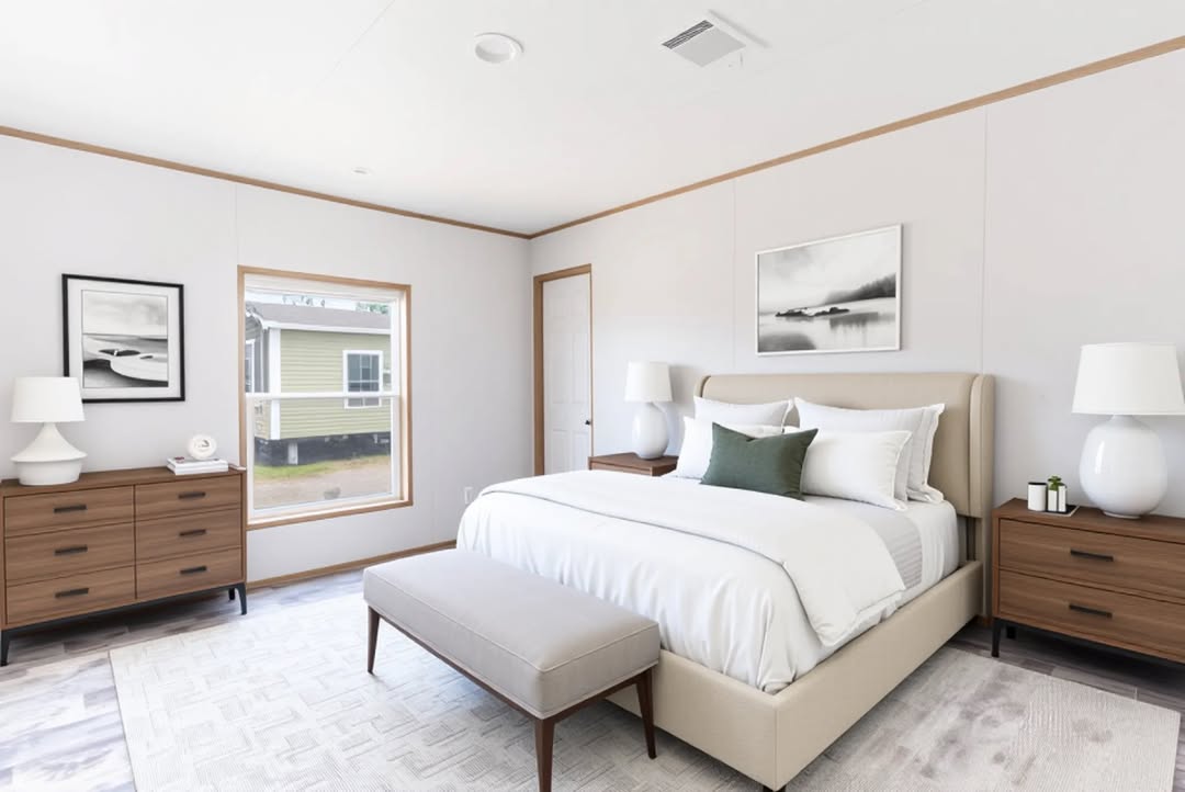 Modern bedroom with a neutral palette, featuring a cozy bed with white linens and green accent pillow. Two wooden nightstands with lamps flank the bed, and a bench sits at the foot. A window with a view and simple artwork create a peaceful atmosphere.