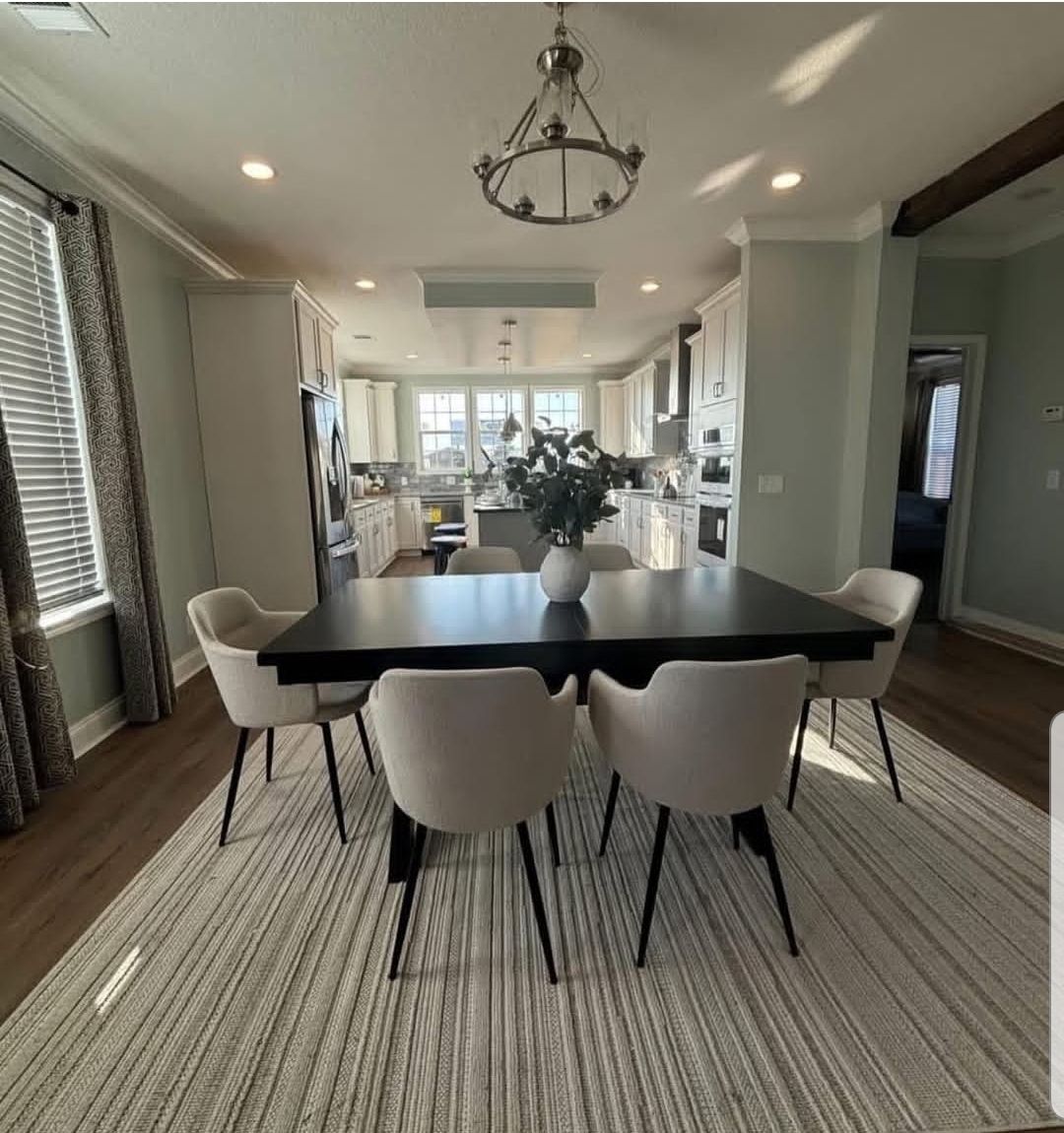 Bright kitchen and dining area with a black table, six cream chairs, and a vase of flowers. White cabinets, large windows, and cozy ambiance.