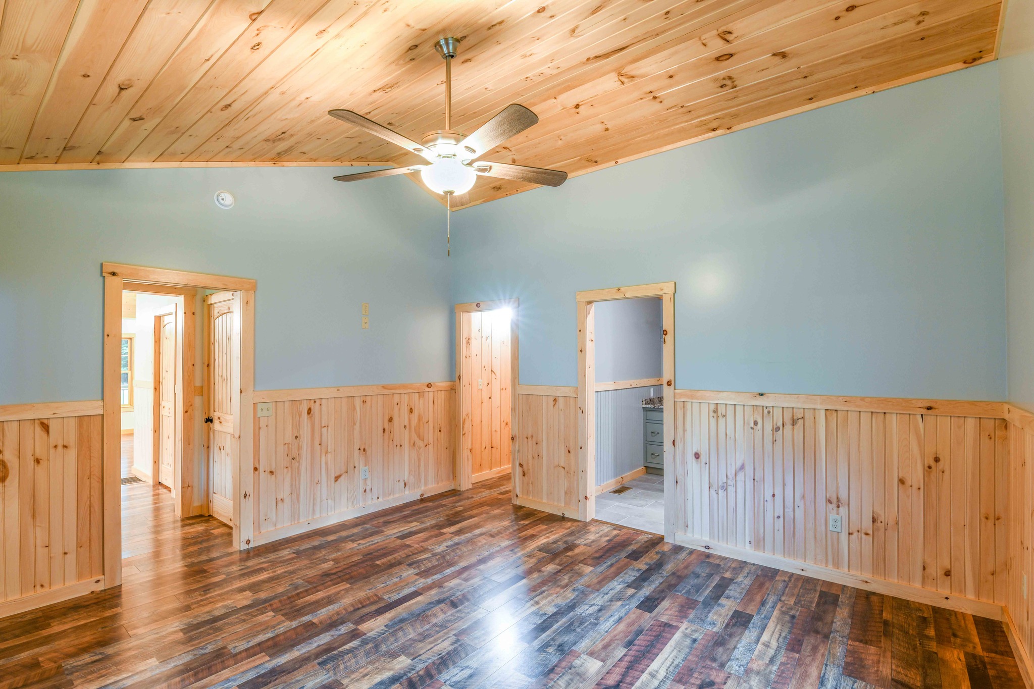 A room with light wood paneling and blue walls features a ceiling fan and polished hardwood floors. Three doorways lead to adjacent rooms. Bright, airy ambiance.