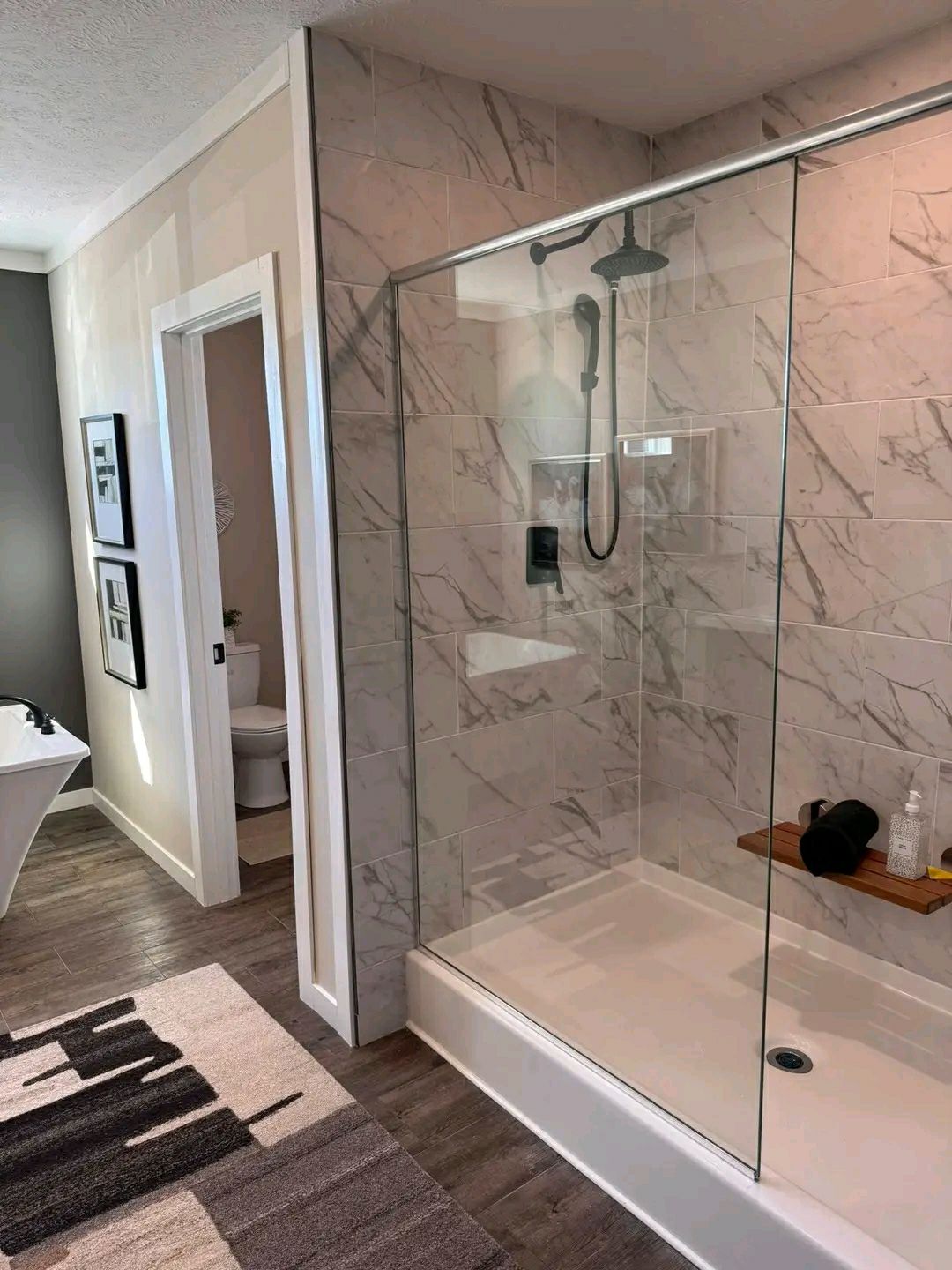 Modern bathroom with a large glass shower featuring marble tiles and a rainfall showerhead. Adjacent is a wooden floor with a patterned rug.