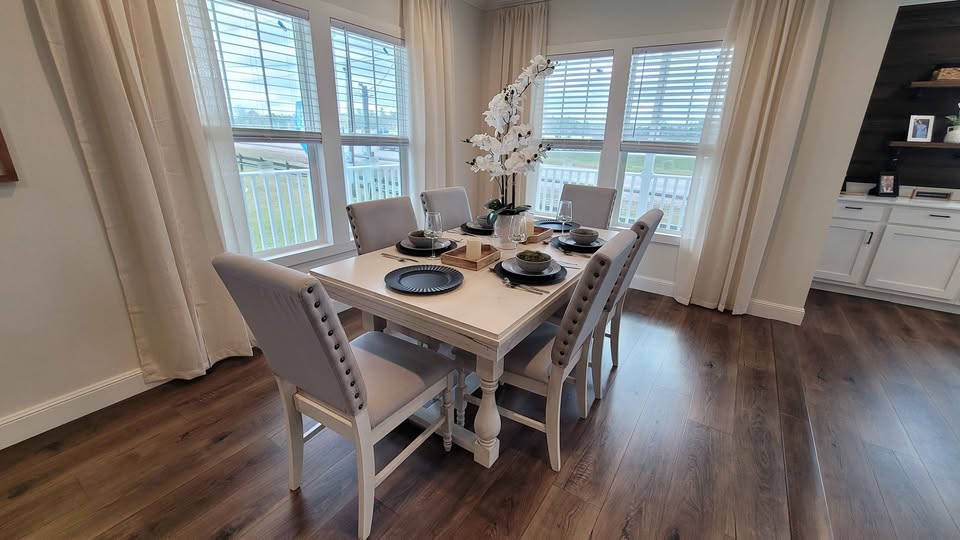 Elegant dining room with a square table set for six, featuring gray upholstered chairs and a white orchid centerpiece, in a bright room with large windows.