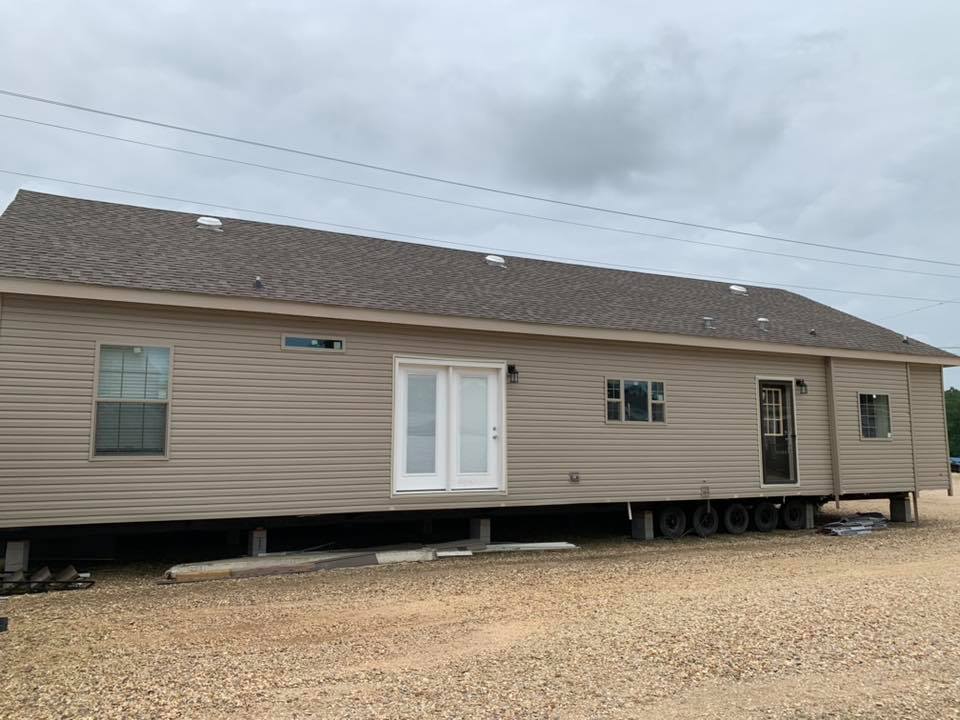 A beige mobile home on wheels sits on a gravel lot under a cloudy sky. It has a shingled roof, white double doors, and multiple windows.