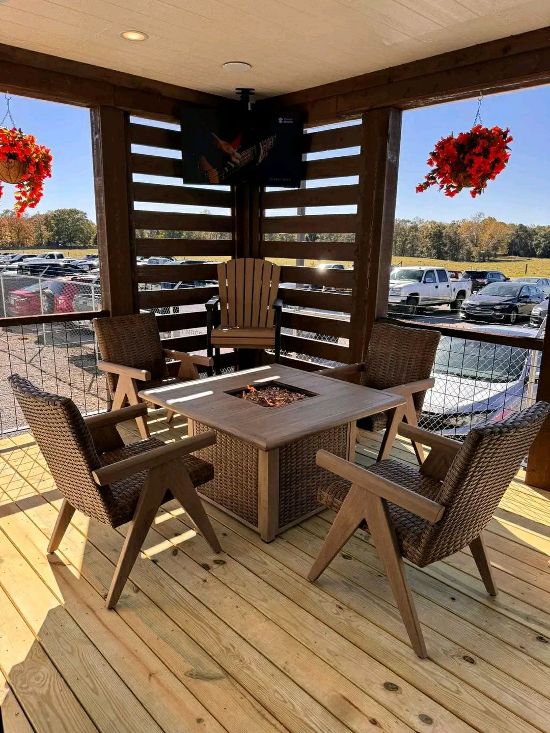 Cozy outdoor patio with four wicker chairs around a square fire pit table. Wooden deck, hanging red flowers, and cars visible in the background.