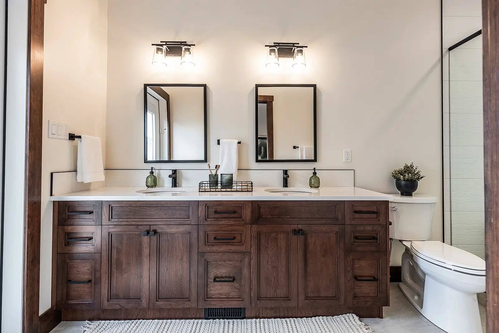 Stylish bathroom with dual wooden vanity, two mirrors, and modern light fixtures. Features white towels, decorative green vases, and a plant by the toilet.